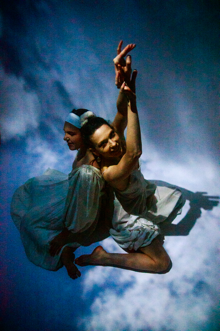Two women sitting in the sky back to back, with one reaching upwards.